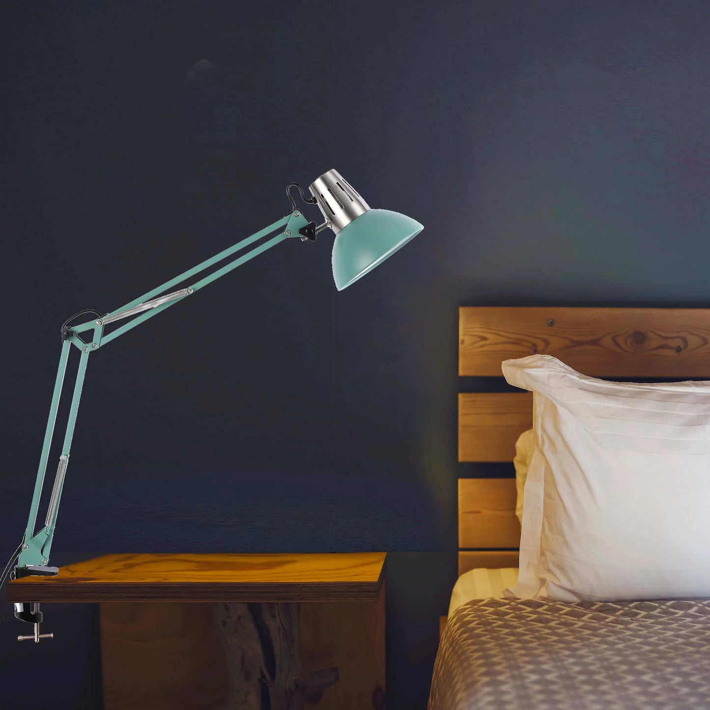 An adjustable, balanced arm desk lamp sits on a wooden bedside table next to a bed. It's providing bright, directional light.