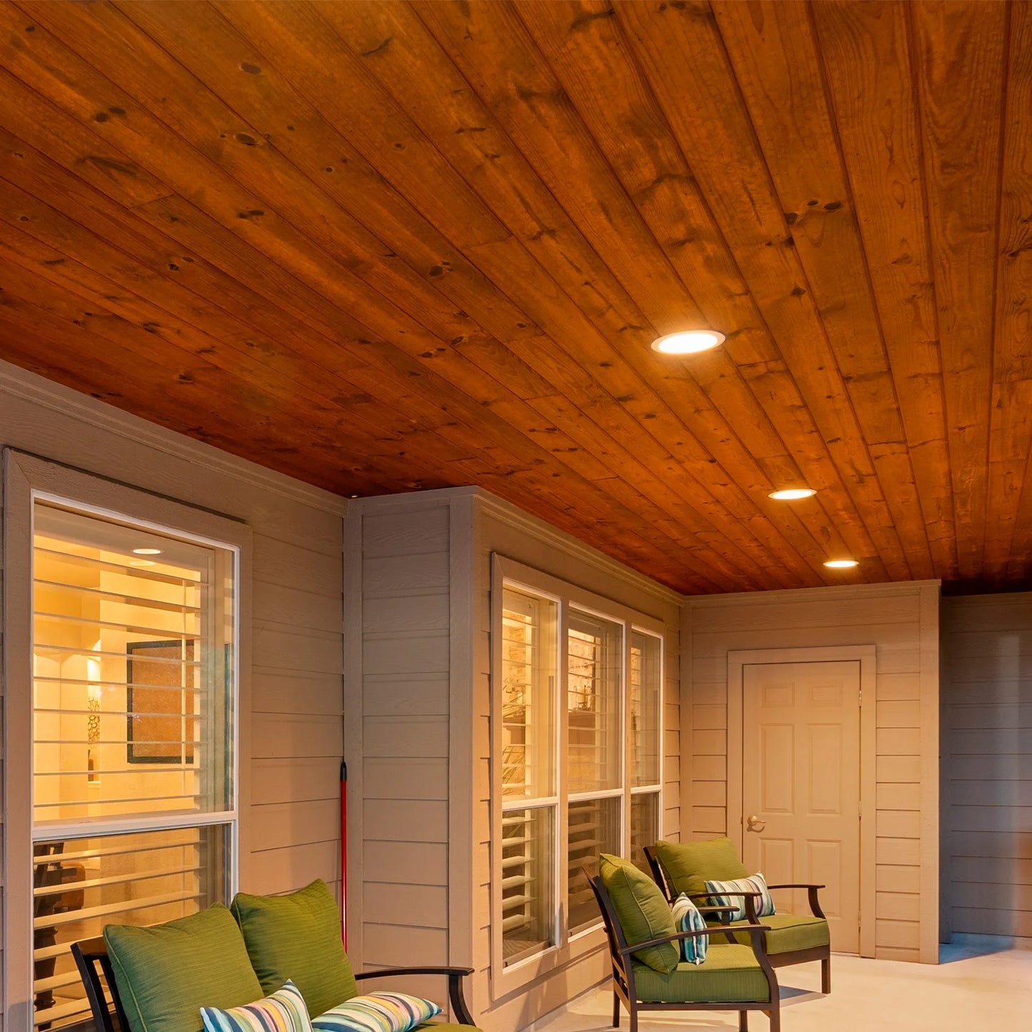 Warm lighting from recessed ceiling lights illuminates an outdoor porch with wooden ceiling and neutral coloured walls.
