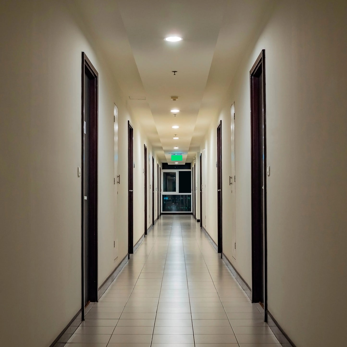 Recessed ceiling lights illuminate a long corridor with doors lining each side, and an exit sign at the end.