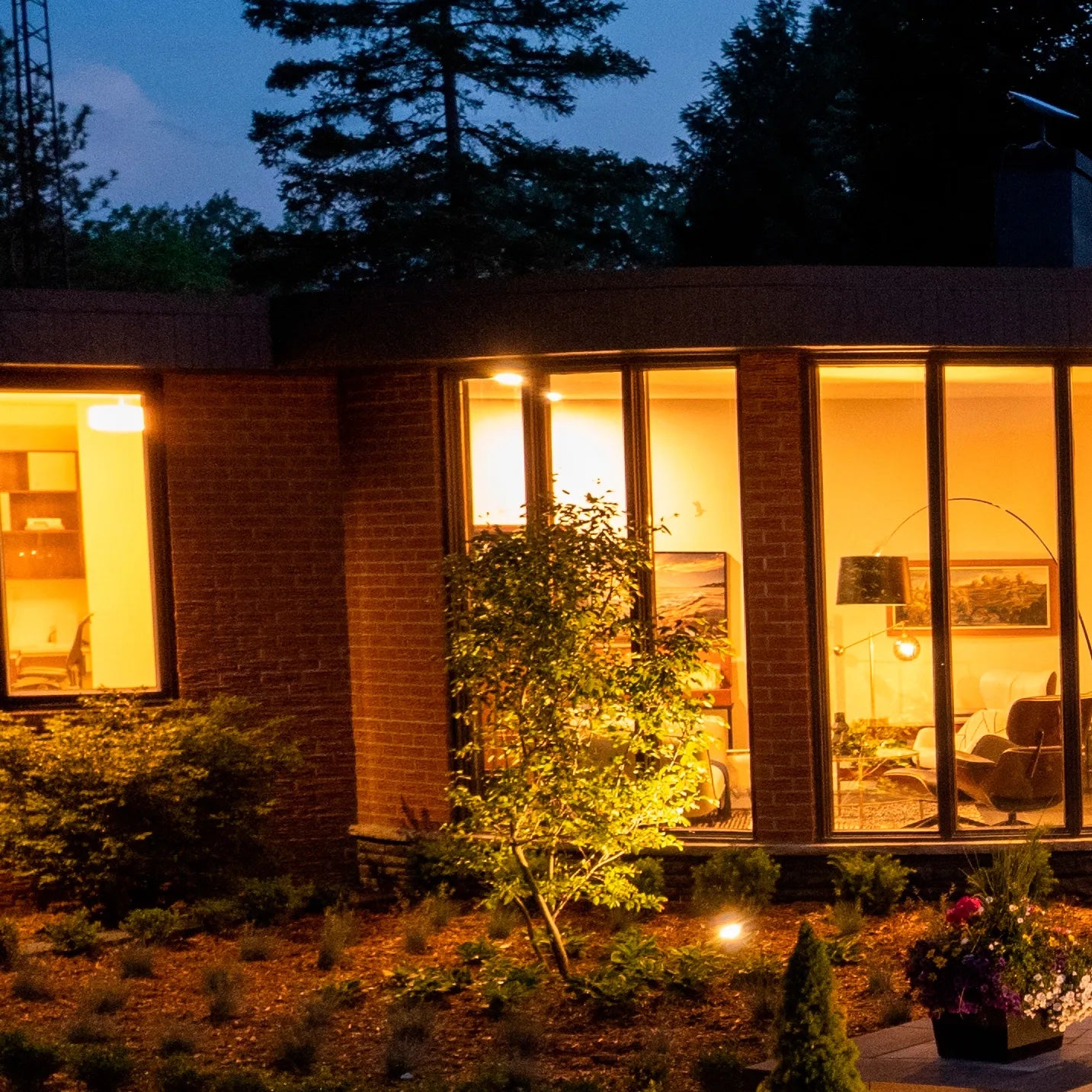 Warm interior lighting shines from a modern house at dusk, illuminating the garden with a welcoming glow.