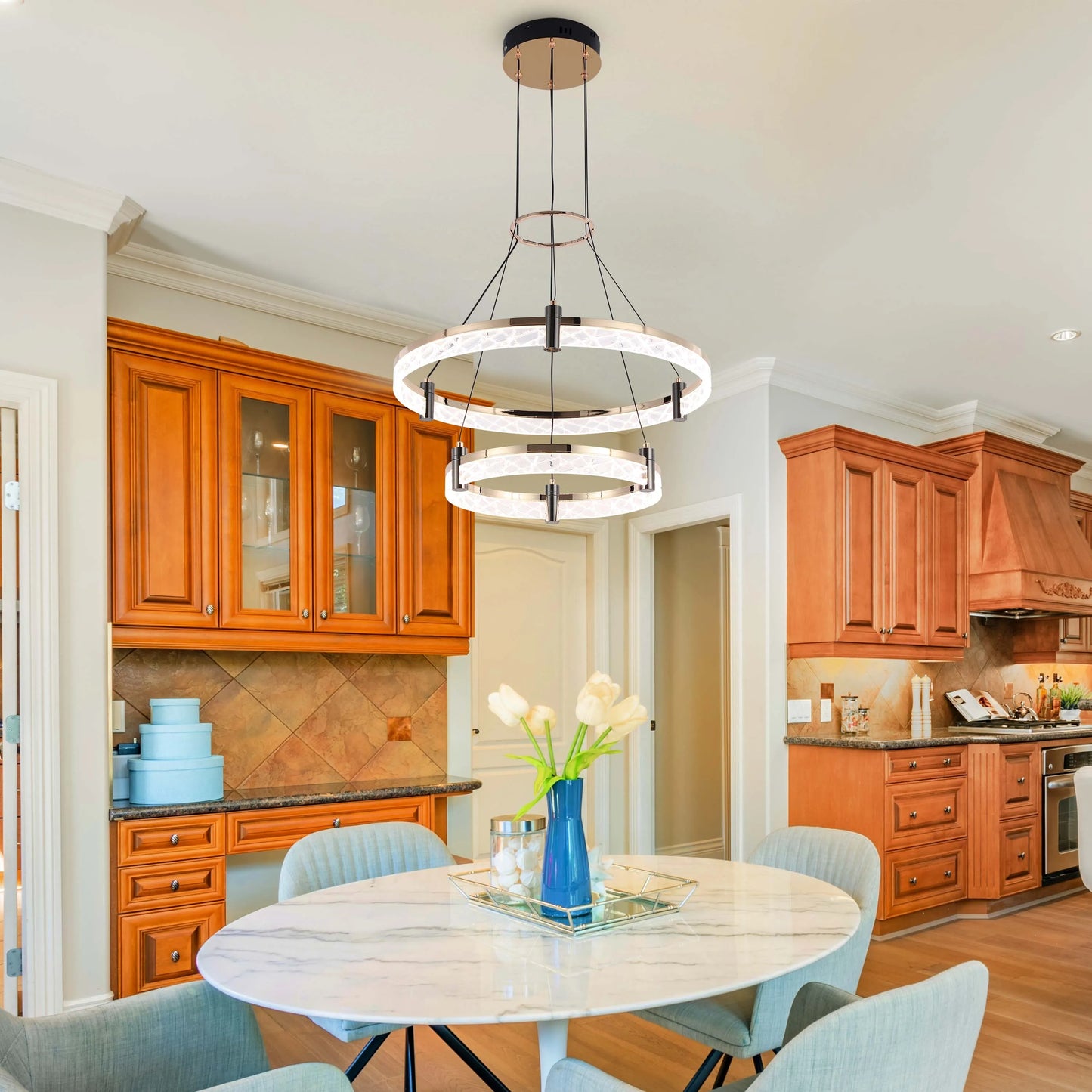 A modern two-tiered LED ring chandelier with a rose gold finish hangs over a round marble dining table in a kitchen setting.