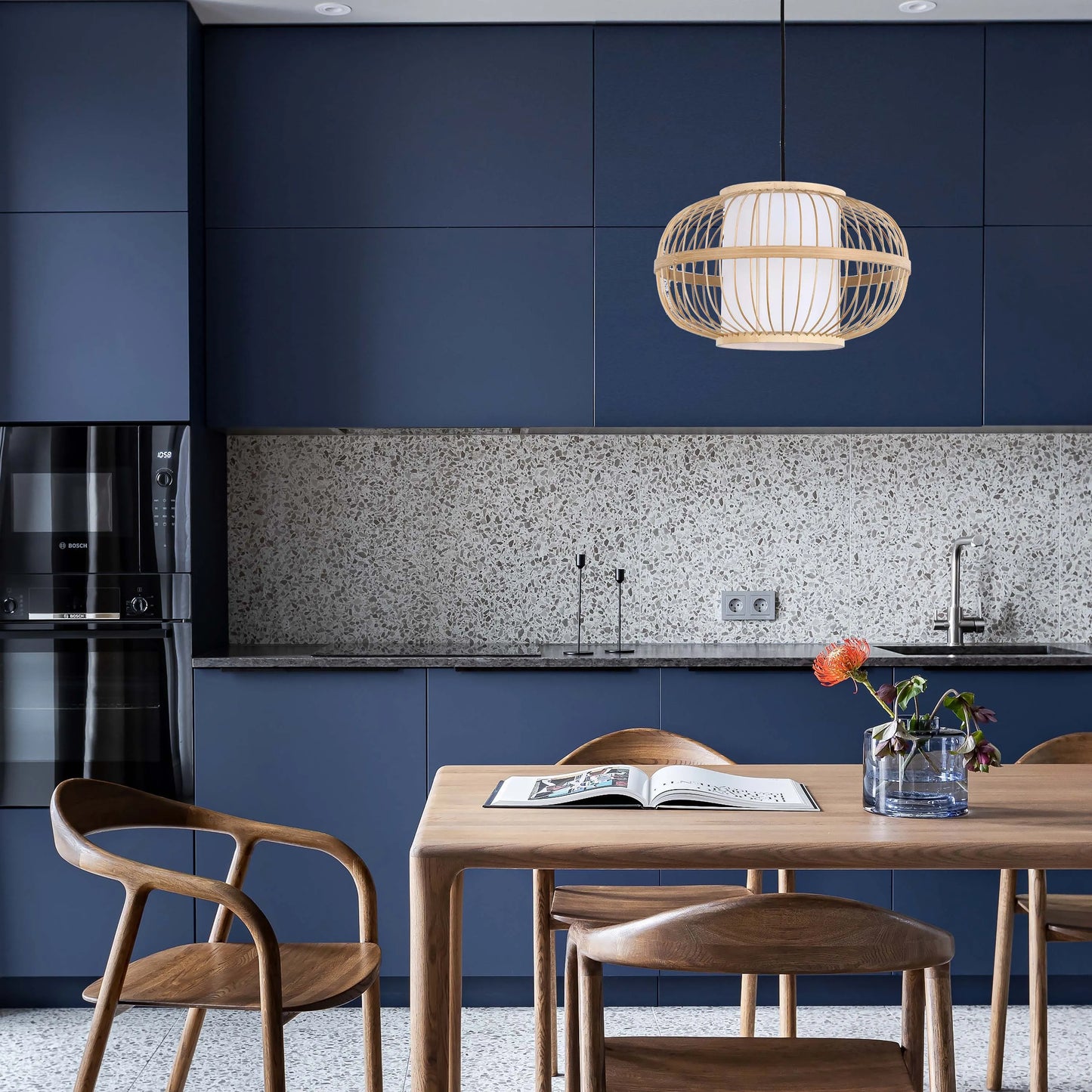 A stylish, woven pendant light hangs above a wooden dining table. Modern blue kitchen cabinets are in the background.