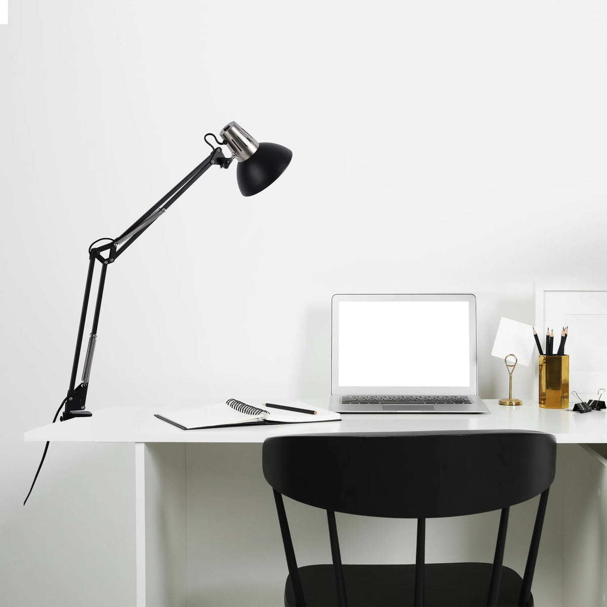 A black adjustable lamp illuminates a minimalist white desk, with a laptop, notebook, and stationery, creating a bright workspace.