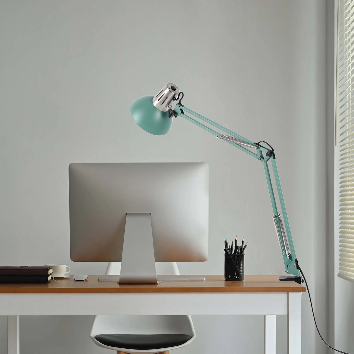 A turquoise adjustable desk lamp illuminates a modern workspace, complemented by natural light filtering through the blinds.