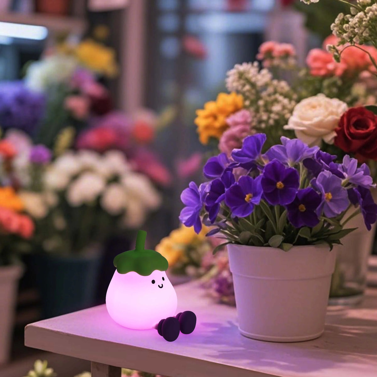 A whimsical, glowing night light shaped like an aubergine, casting a soft, colourful glow amongst a colourful flower display.