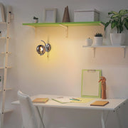 A warm, glowing wall light fixture illuminates a minimalist workspace with white furniture and vibrant green shelves.