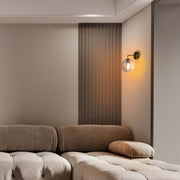 A wall mounted light fixture emits a warm glow from a clear globe. The base is a metallic bronze.