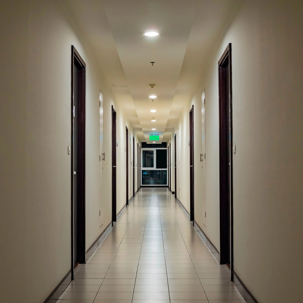 Recessed ceiling lights illuminate a long corridor with doors lining each side, and an exit sign at the end.