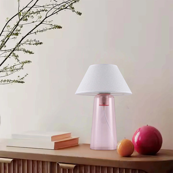 A light pink table lamp with a white lampshade sits on a wooden table, next to fruit and books. A tree branch adds a natural touch.