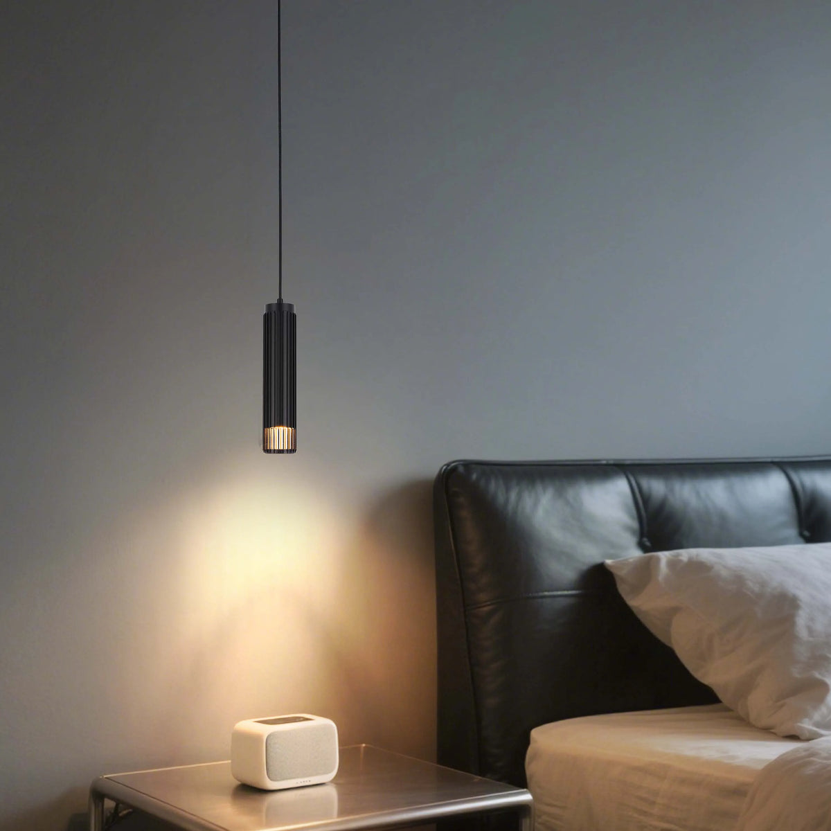 A modern black pendant light casts a warm glow in a bedroom setting, illuminating a bedside table and adjacent bed.