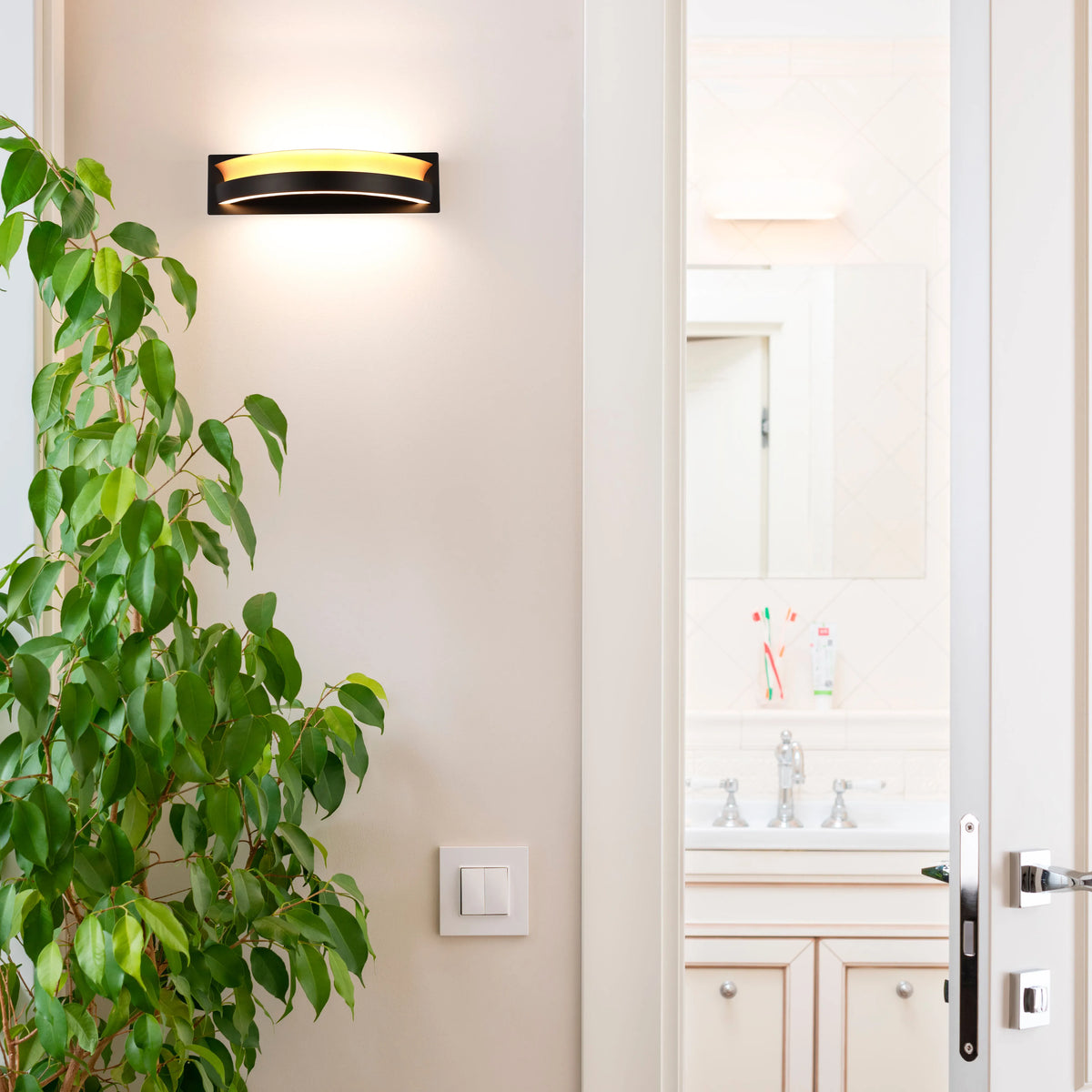 A contemporary wall light with a black surround emits a warm glow. Positioned above a light switch on a cream-coloured wall.