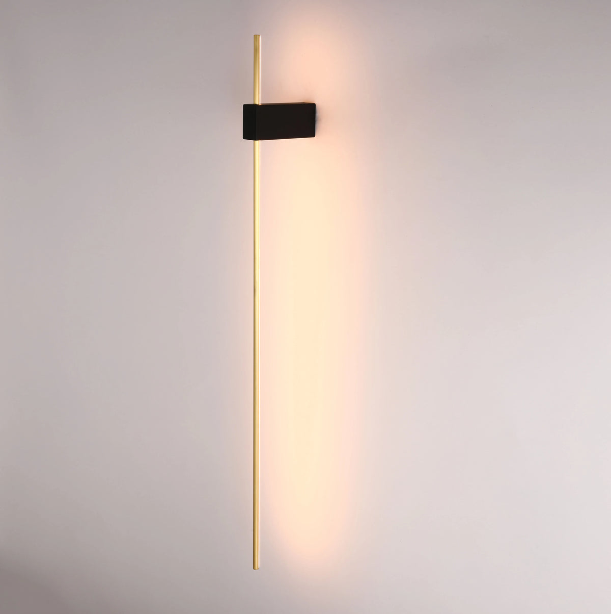 Sleek, modern wall light with a long gold bar and a black rectangular mount, casting a warm glow onto the wall.