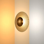 A gold wall light with a round, textured shade, evenly lit with a soft, diffused glow.