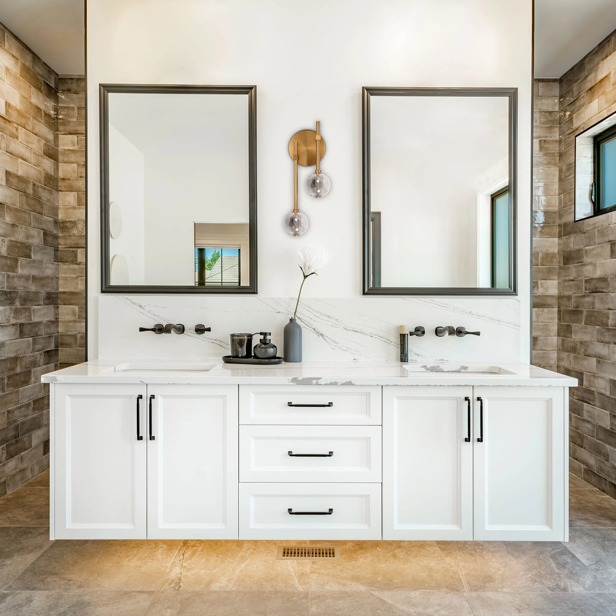 A pair of gold wall sconces with clear glass globe shades above a white vanity unit in a bathroom with brick-effect tiles.