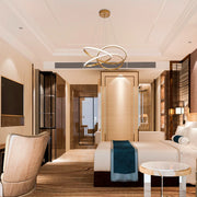 A modern statement LED chandelier with entwined golden rings, suspended above a hotel bedroom's bed.