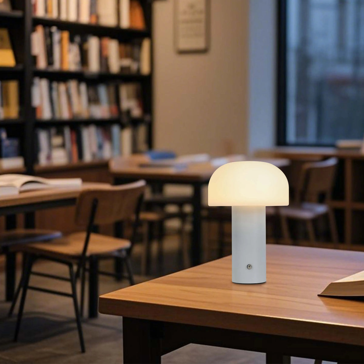 A modern mushroom table lamp emitting a warm glow, set on a wooden table in a library setting, creating a cosy atmosphere.