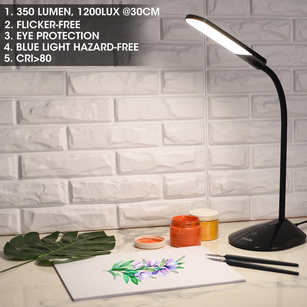 A black desk lamp illuminates art supplies and a watercolour painting on a white desk, with a brick effect wall in the background.
